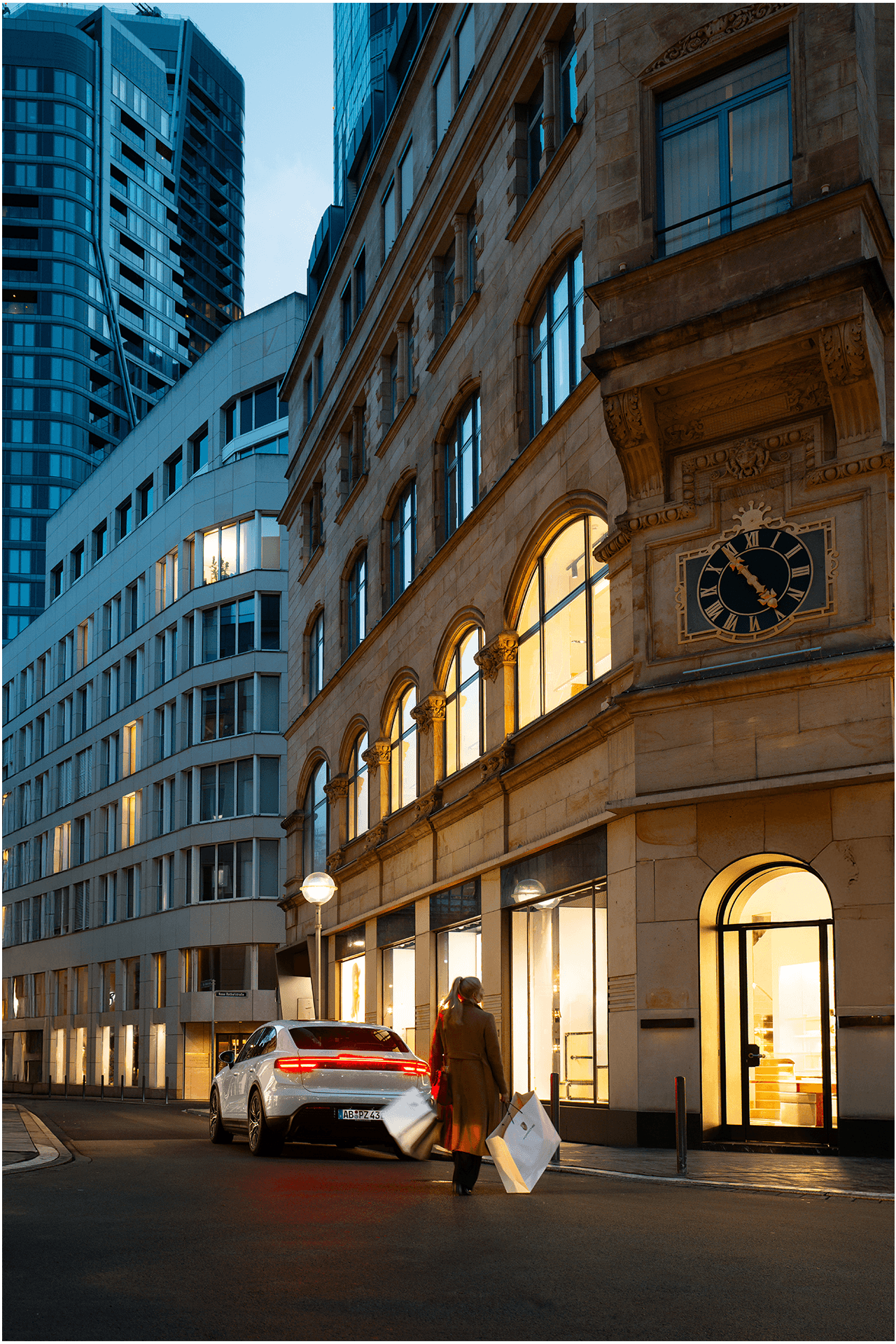 Eine Frau geht in der abendlichen Stadt mit zwei Einkaufstaschen in der Hand auf einen geparkten Macan 4 zu